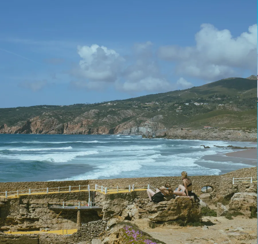 Guincho Beach with rocky cliffs, blue waves and two people sitting overlooking the sea.