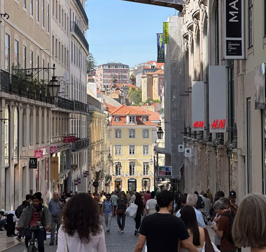 A busy pedestrian street with historic buildings, shops and people walking and cycling.
