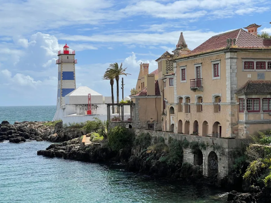 A coastal scene with a striped lighthouse, a historic beige building, palm trees and clear blue water